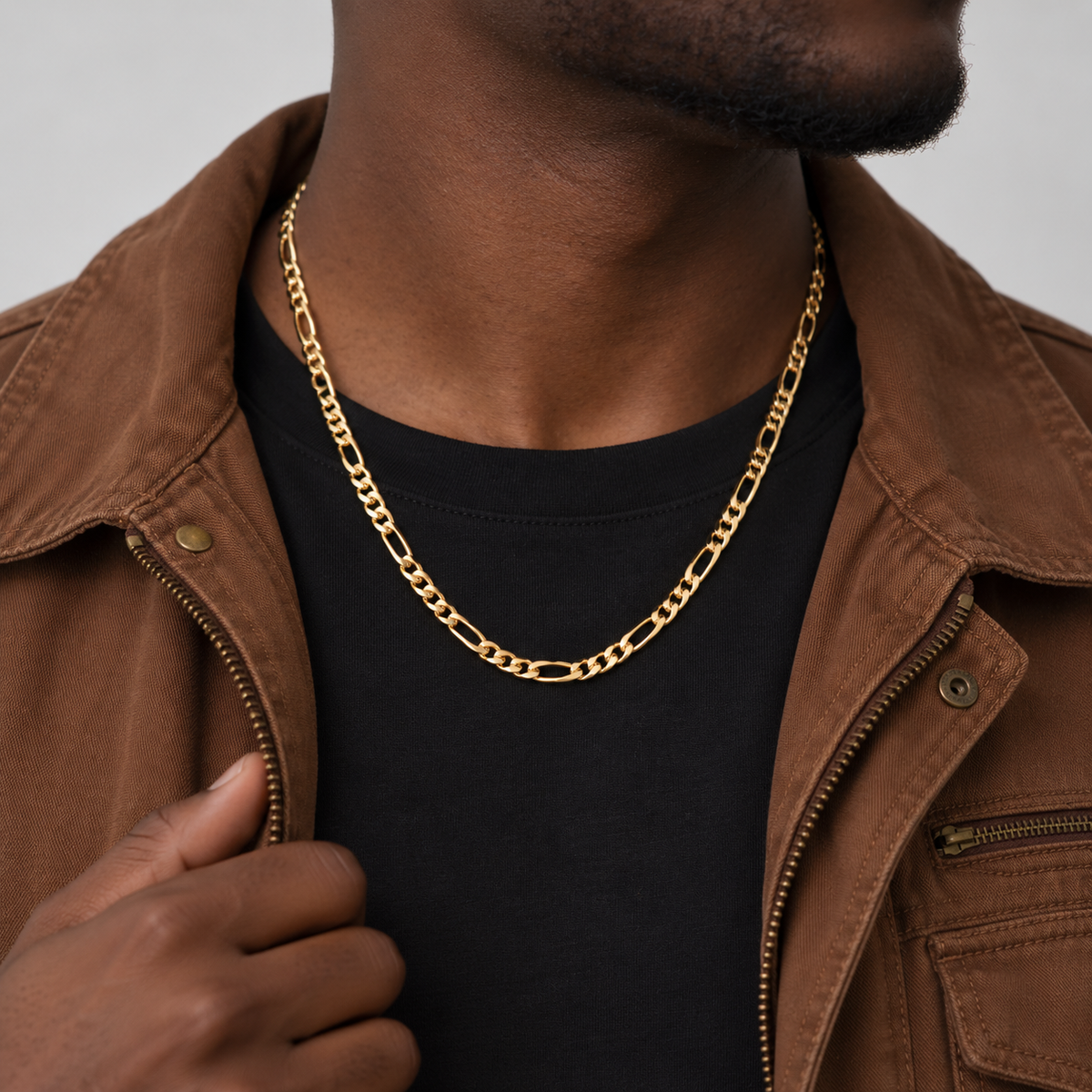 Unveiled 5mm Figaro chain in 18k PVD gold, 50cm, worn by the Unveiled model in a brown canvas jacket