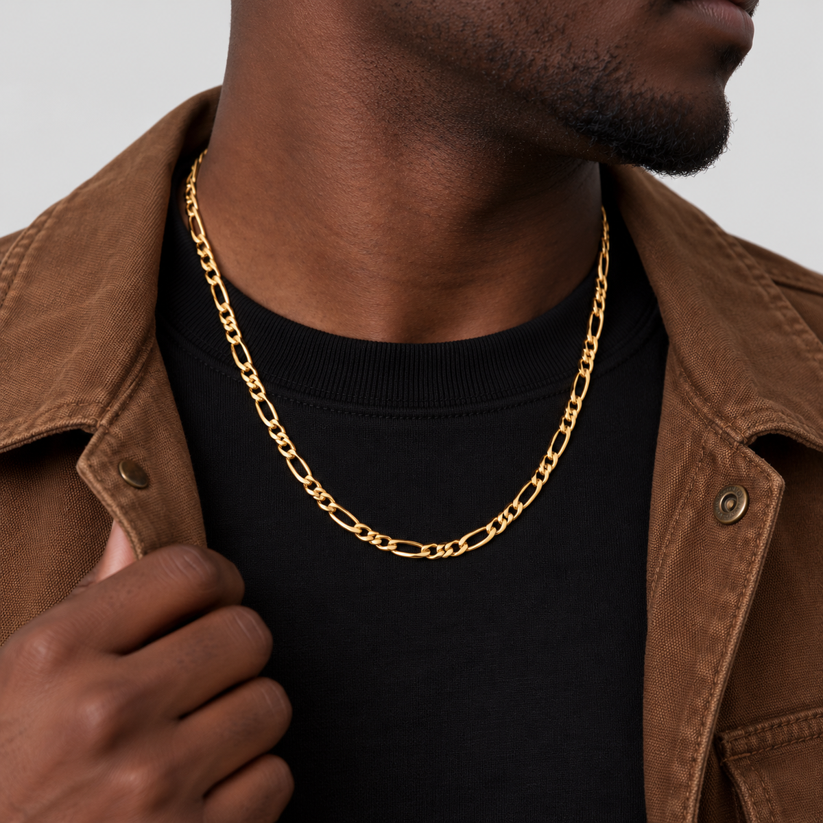 Unveiled 5mm Figaro chain in 18k PVD gold, 50cm, worn by the Unveiled model in a brown canvas jacket, looking away