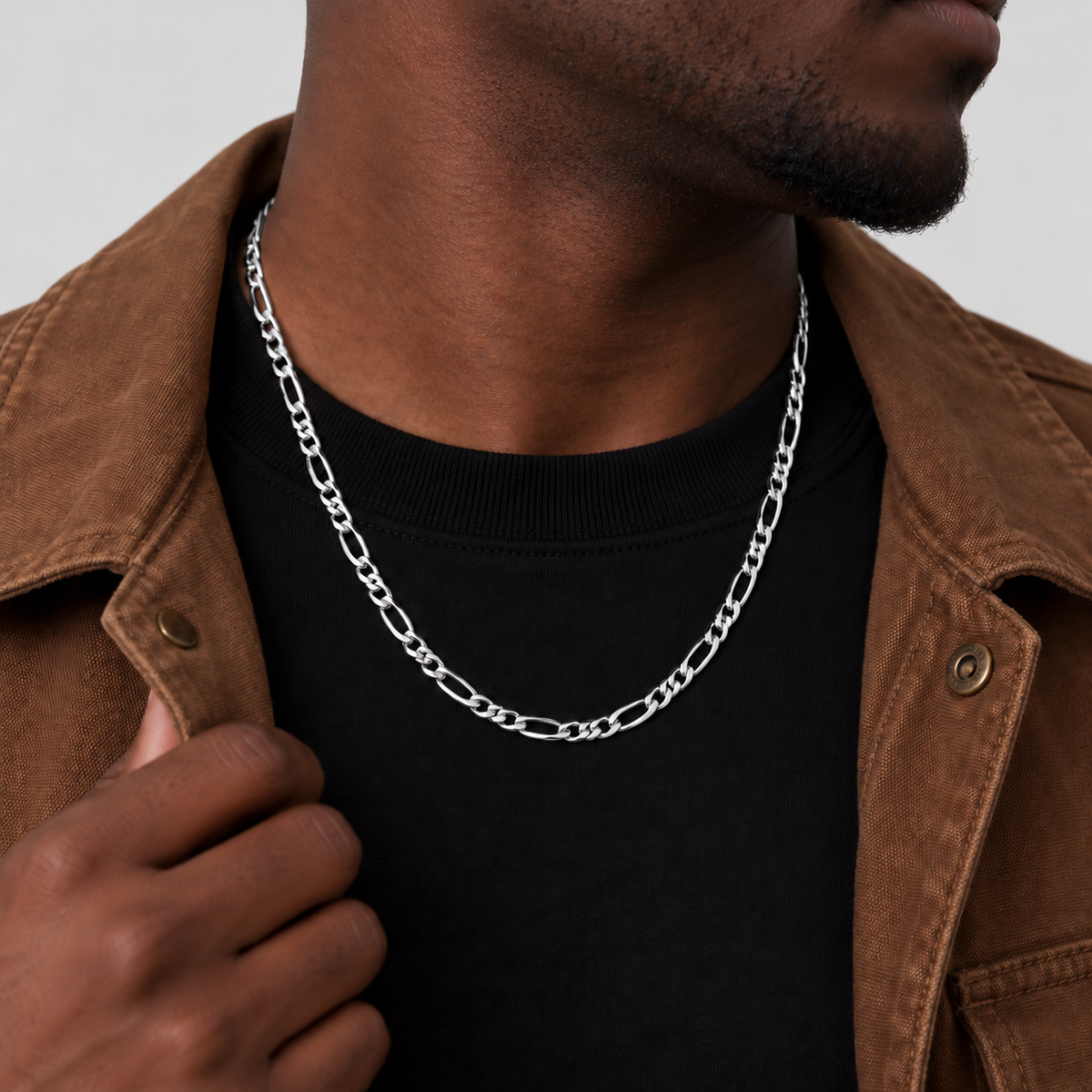 Unveiled 5mm Figaro chain in stainless steel silver, 50cm, worn by the Unveiled model in a brown canvas jacket, looking away