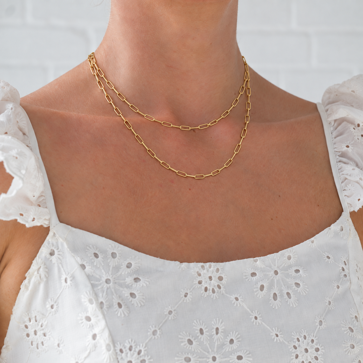 Two gold paperclip necklaces layered on a model wearing a white embroidered dress